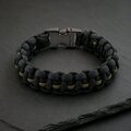 A black and olive green braided paracord bracelet with a gunmetal buckle resting on a dark slate surface.