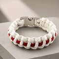 A braided white and red paracord bracelet with a silver metal buckle, resting on a textured gray surface.