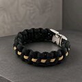 An angled top-down view of the black and gold paracord bracelet with a connected silver buckle on a gray textured background.