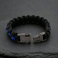 The paracord survival bracelet is unbuckled, displaying the two halves of the dark metal clasp against a textured stone backdrop.