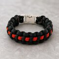 A black and bright orange paracord bracelet with a silver side-release buckle, lying on a light grey textured surface.