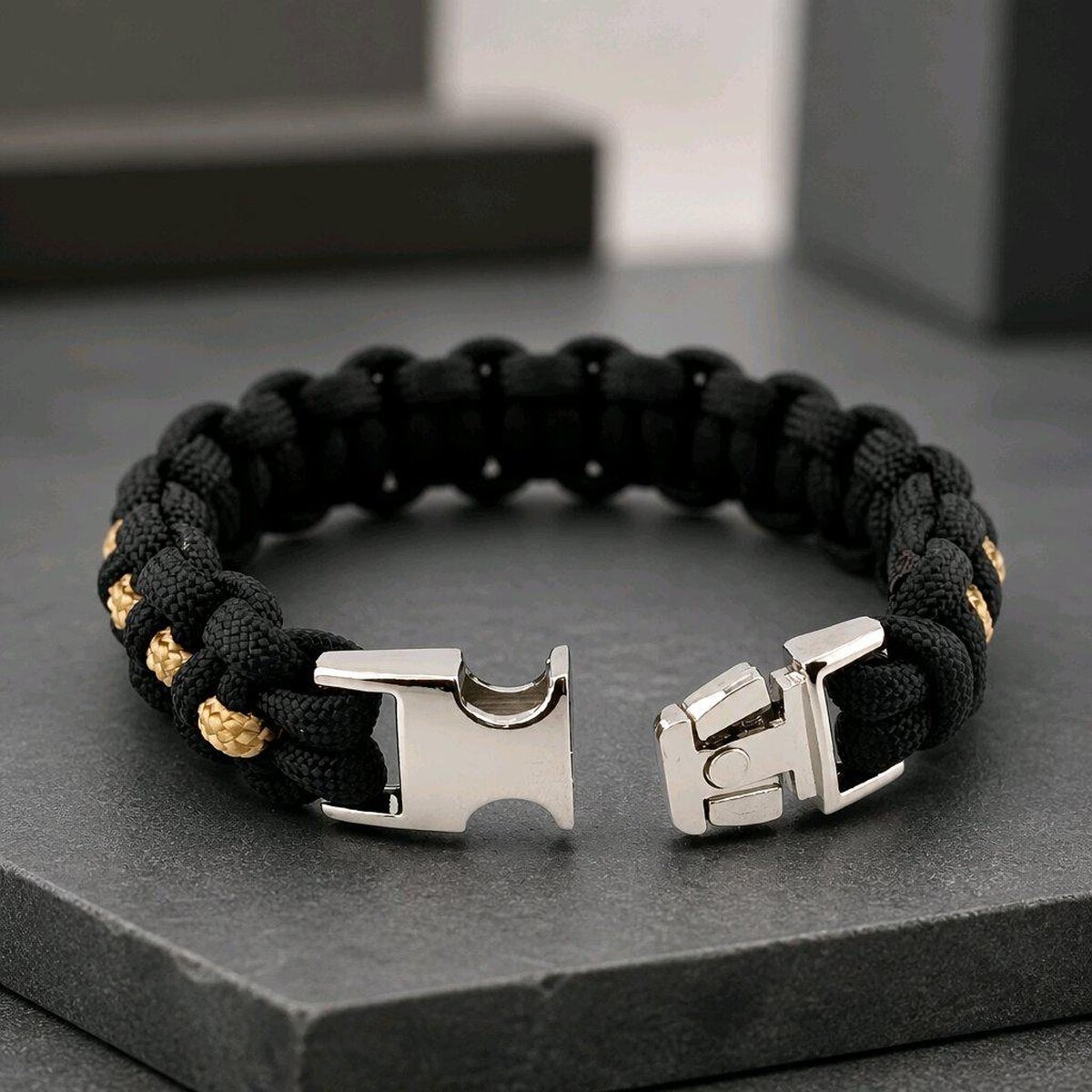 The black and gold paracord bracelet with its metal side-release buckle unlatched and resting open on a dark stone tile.