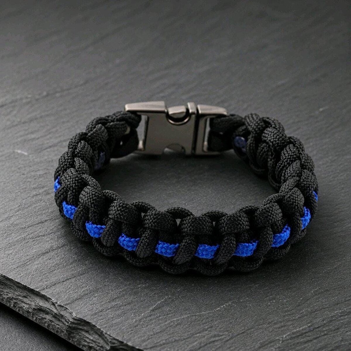 A black and blue paracord survival bracelet rests on a dark slate surface, showing the metal side-release buckle.