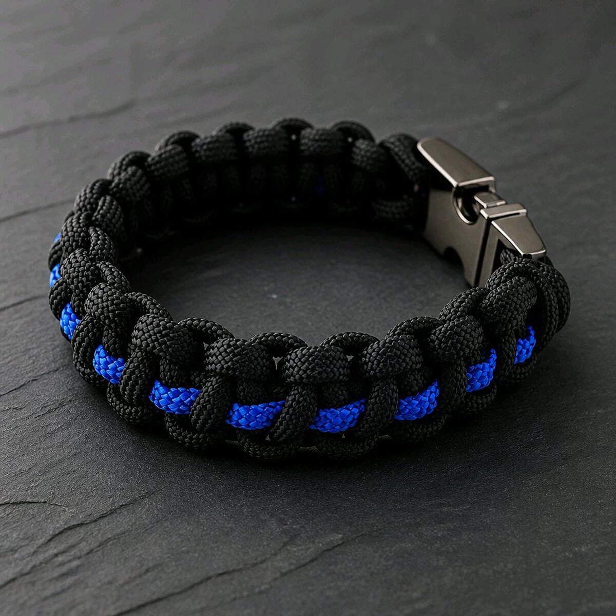 The black braided paracord bracelet with a thin blue center line lies flat on a dark stone background.