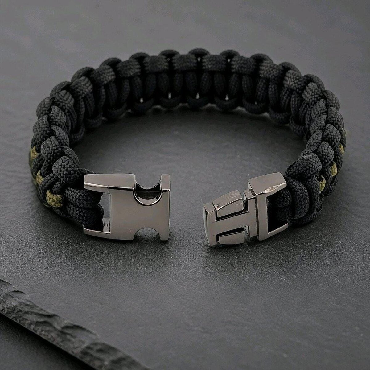 The paracord bracelet with its gunmetal buckle unclasped on a dark stone surface.