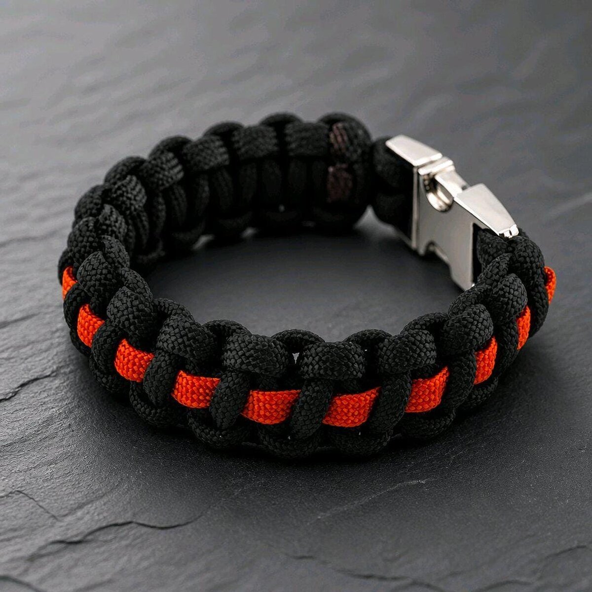A braided black and orange paracord bracelet featuring a metal buckle, presented on a dark slate surface.