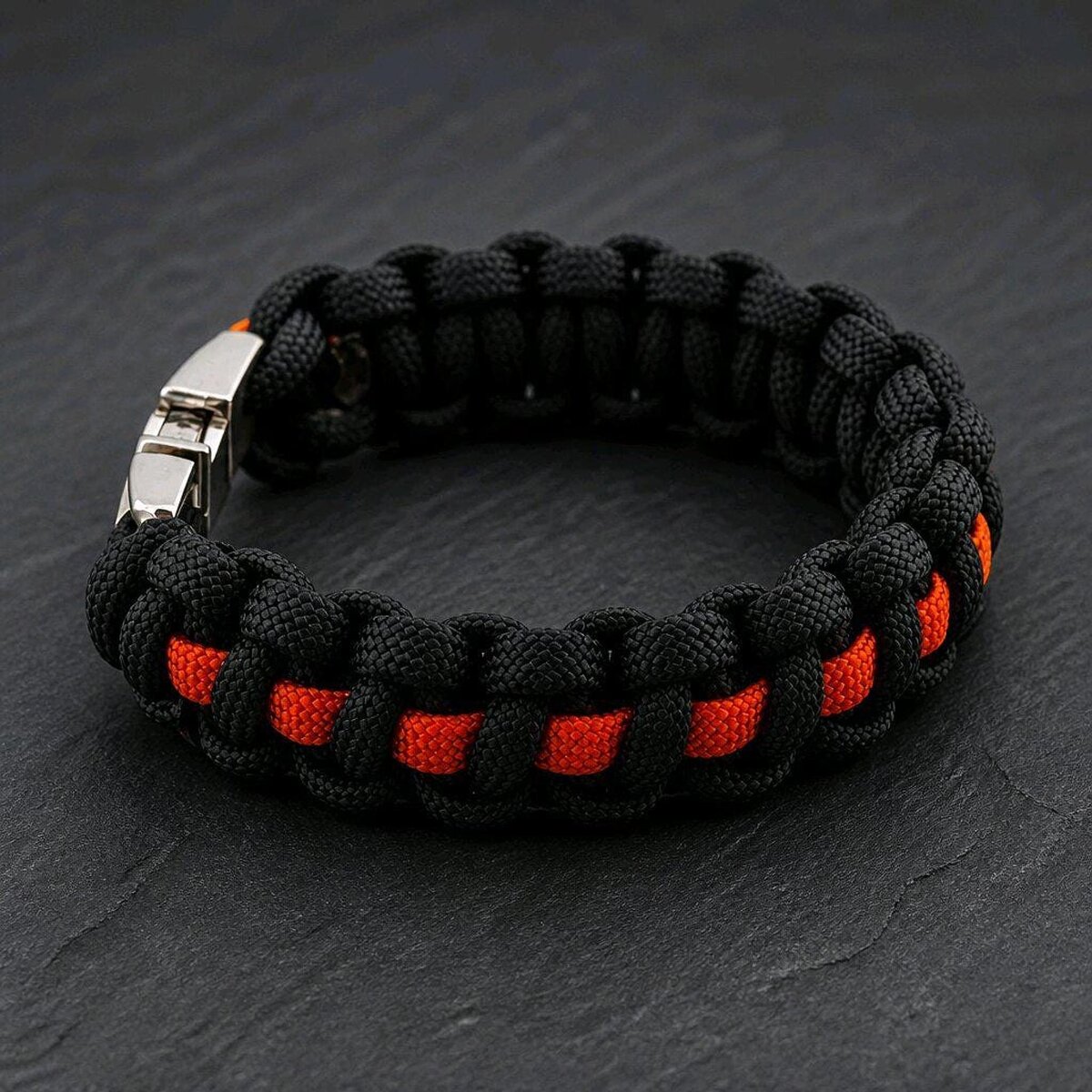 A thick paracord bracelet made of black cord with a thin orange center line, fastened with a silver buckle on a dark background.