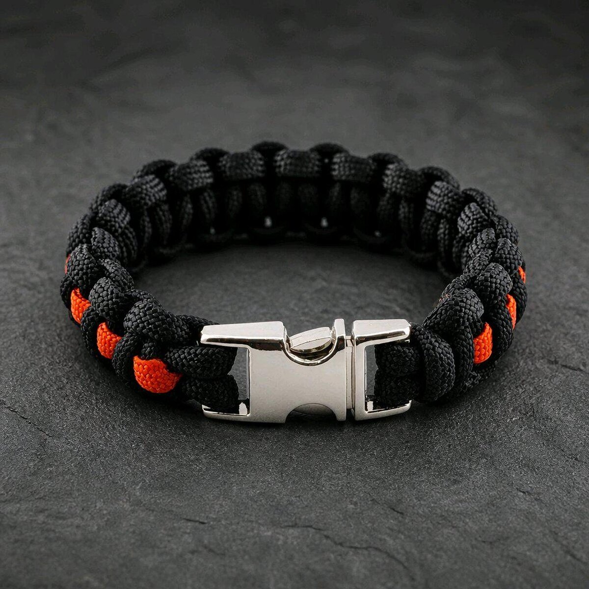A survival bracelet woven from black and orange paracord, shown closed with its polished silver plastic-style side-release buckle on a dark stone background.
