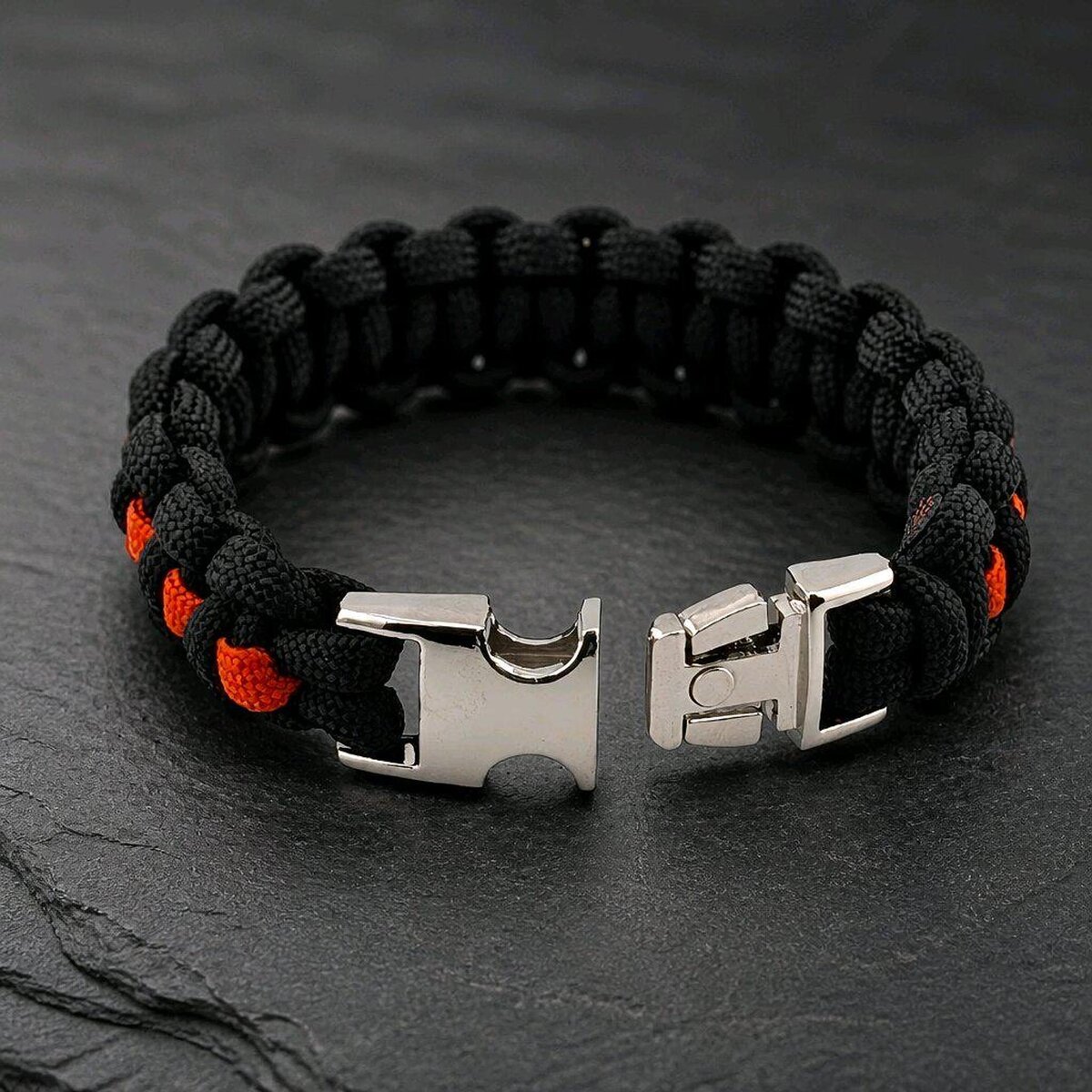 A black and orange woven paracord bracelet displayed with its silver side-release buckle unlatched on a slate backdrop.