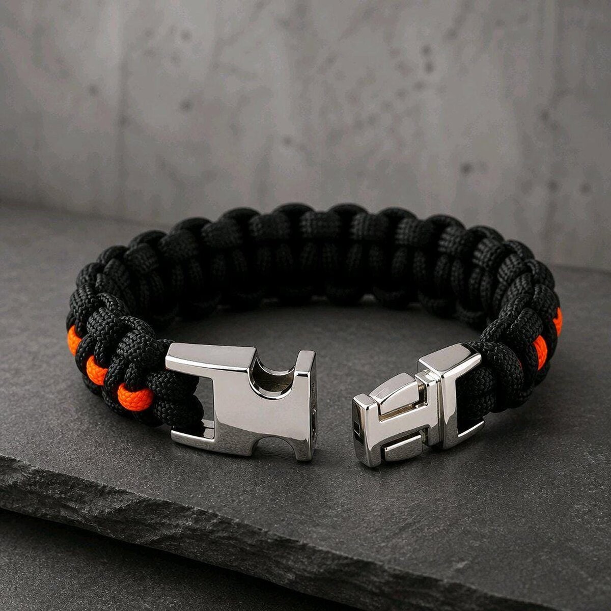 The paracord bracelet with its metal side-release buckle unlatched, resting on textured stone.