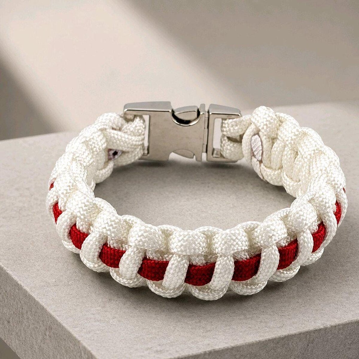 A braided white and red paracord bracelet with a silver metal buckle, resting on a textured gray surface.