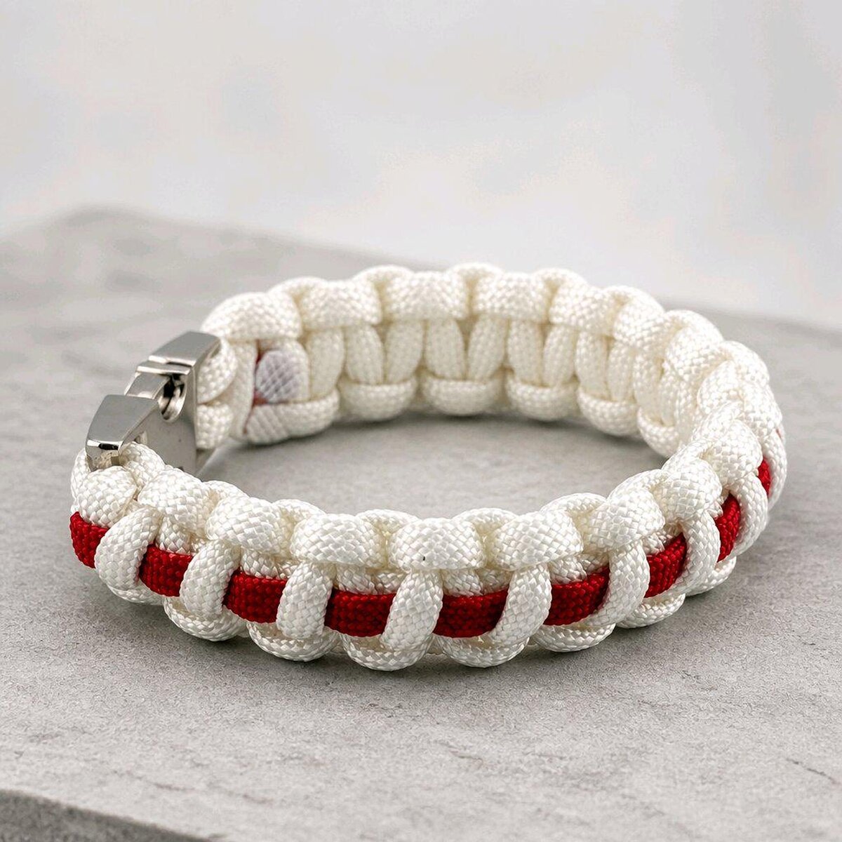 A white and red cobra-knot paracord bracelet viewed from the side on a concrete background.