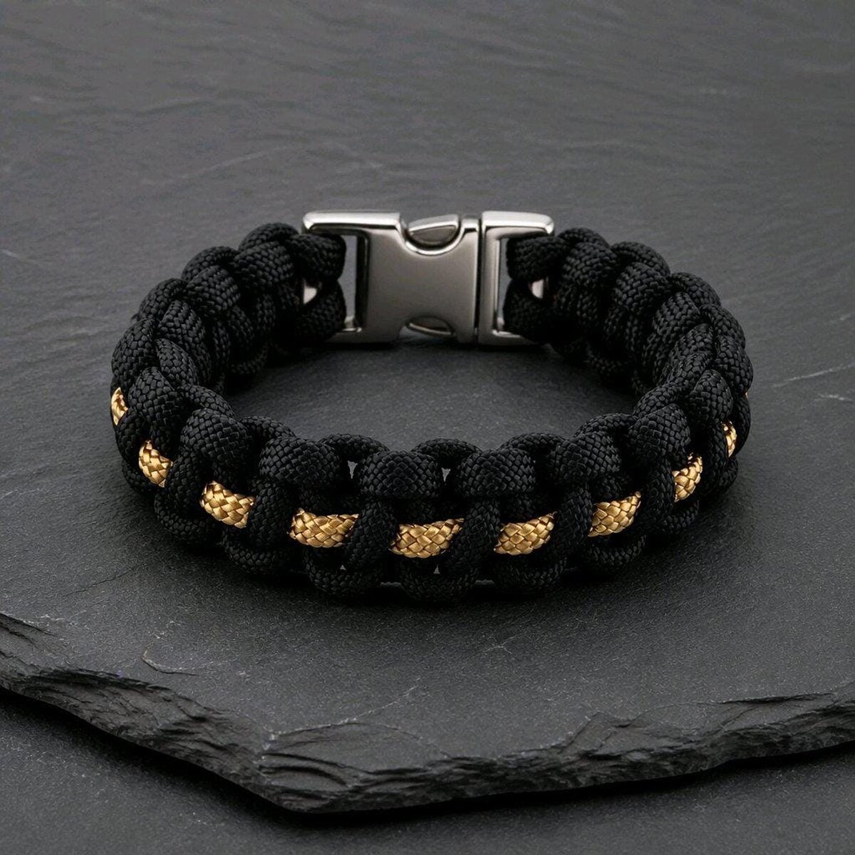 A black and gold paracord bracelet with a silver side-release buckle resting on a dark slate surface.