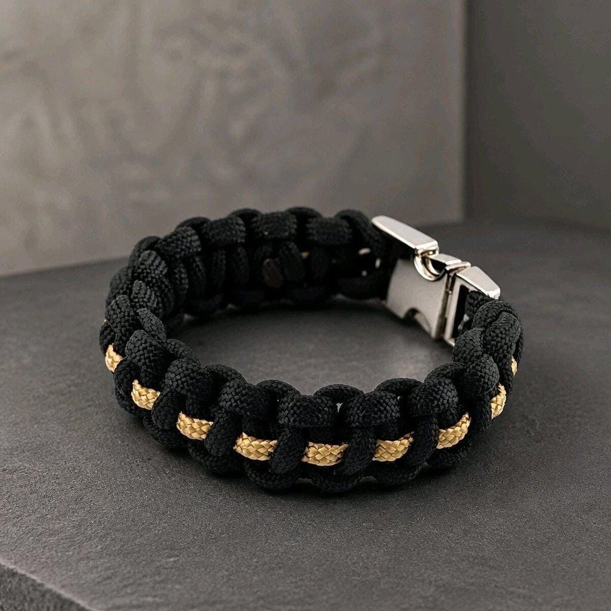 An angled top-down view of the black and gold paracord bracelet with a connected silver buckle on a gray textured background.