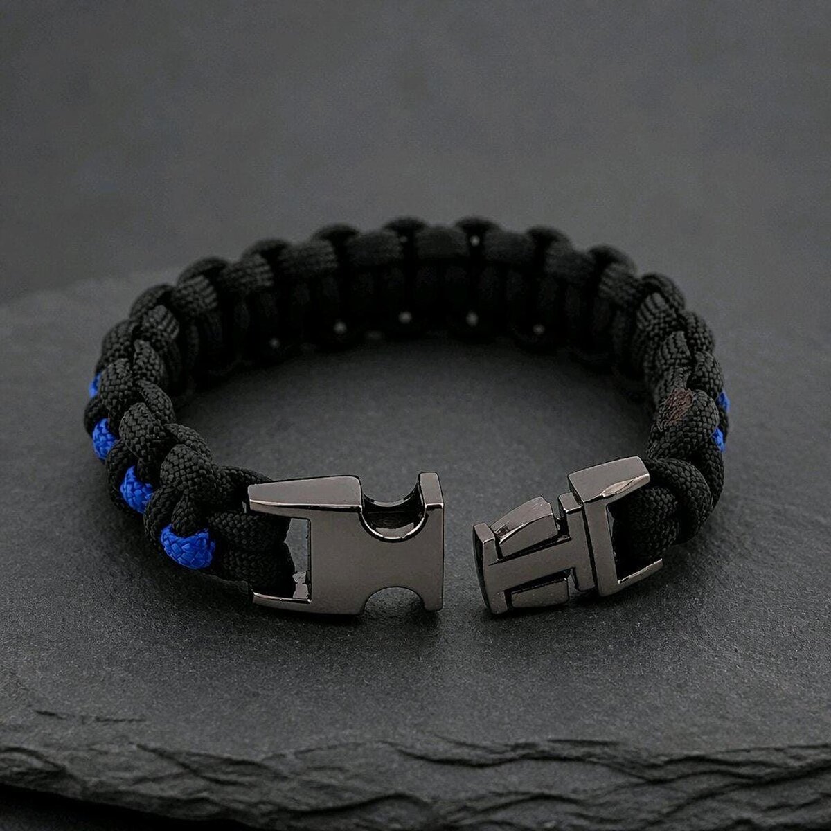 The paracord survival bracelet is unbuckled, displaying the two halves of the dark metal clasp against a textured stone backdrop.