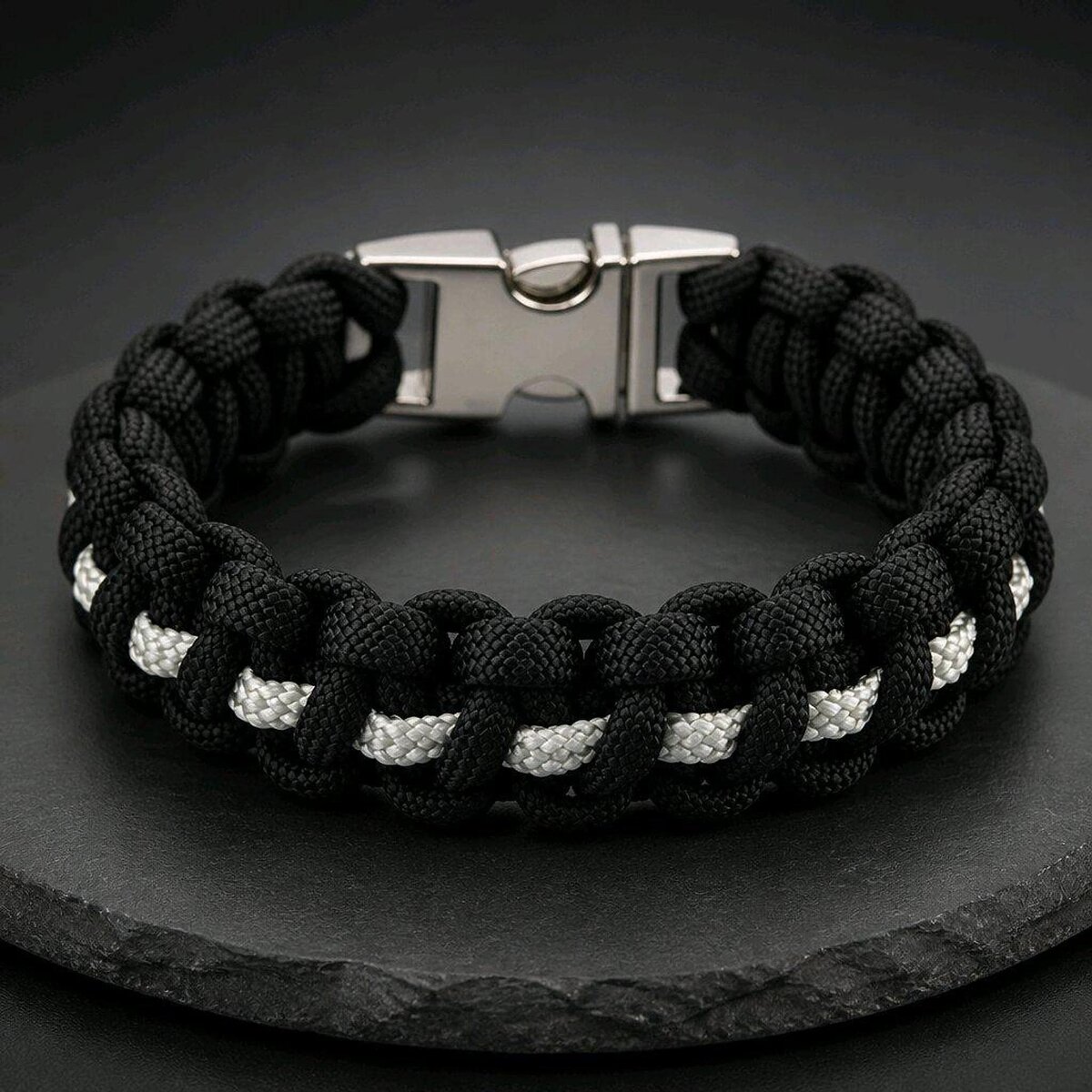 A black and white paracord bracelet with a silver side-release buckle resting on a dark slate surface, viewed from the top with the buckle centered at the back.