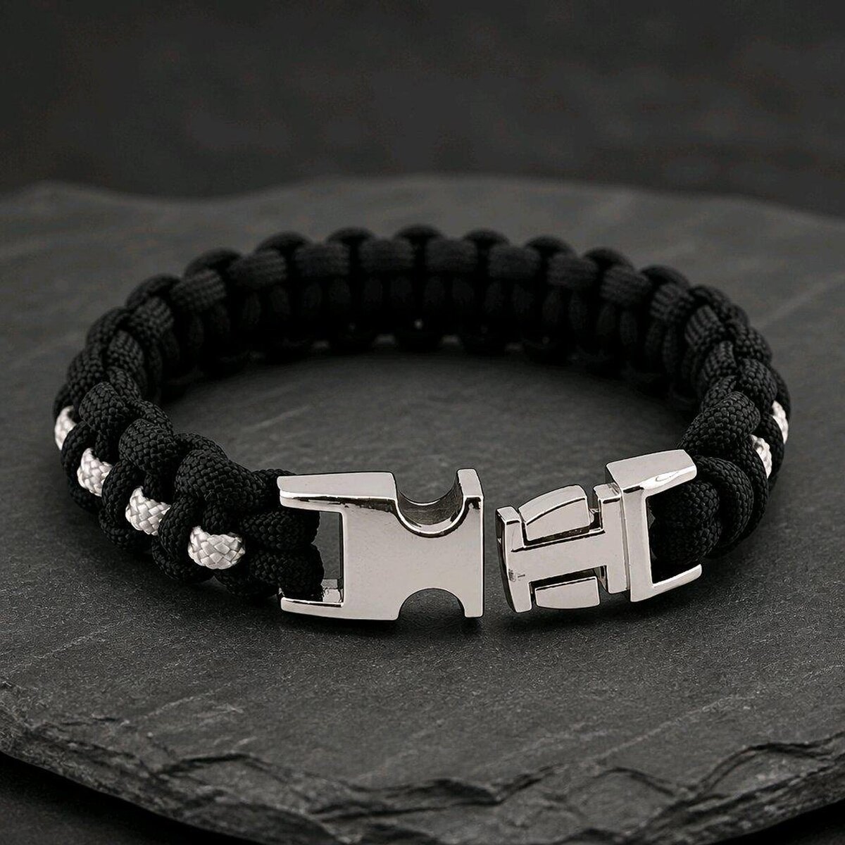 The paracord bracelet displayed with its silver metal buckle unlatched on a dark textured slate background.