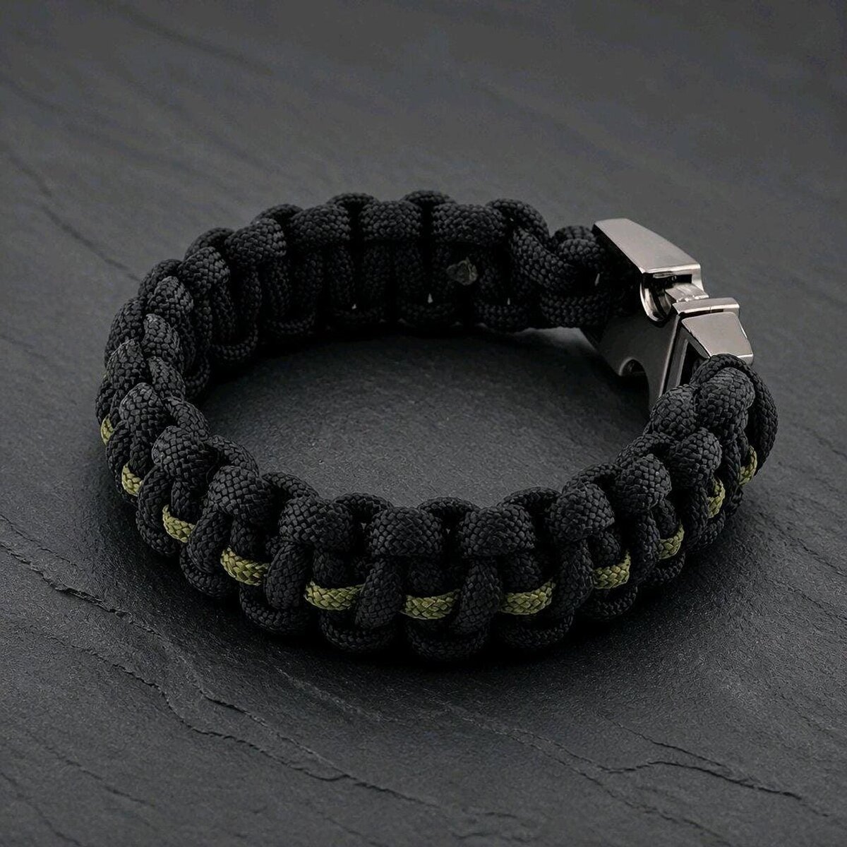 The black paracord bracelet angled on the slate surface, showing the interwoven olive green center stripe.