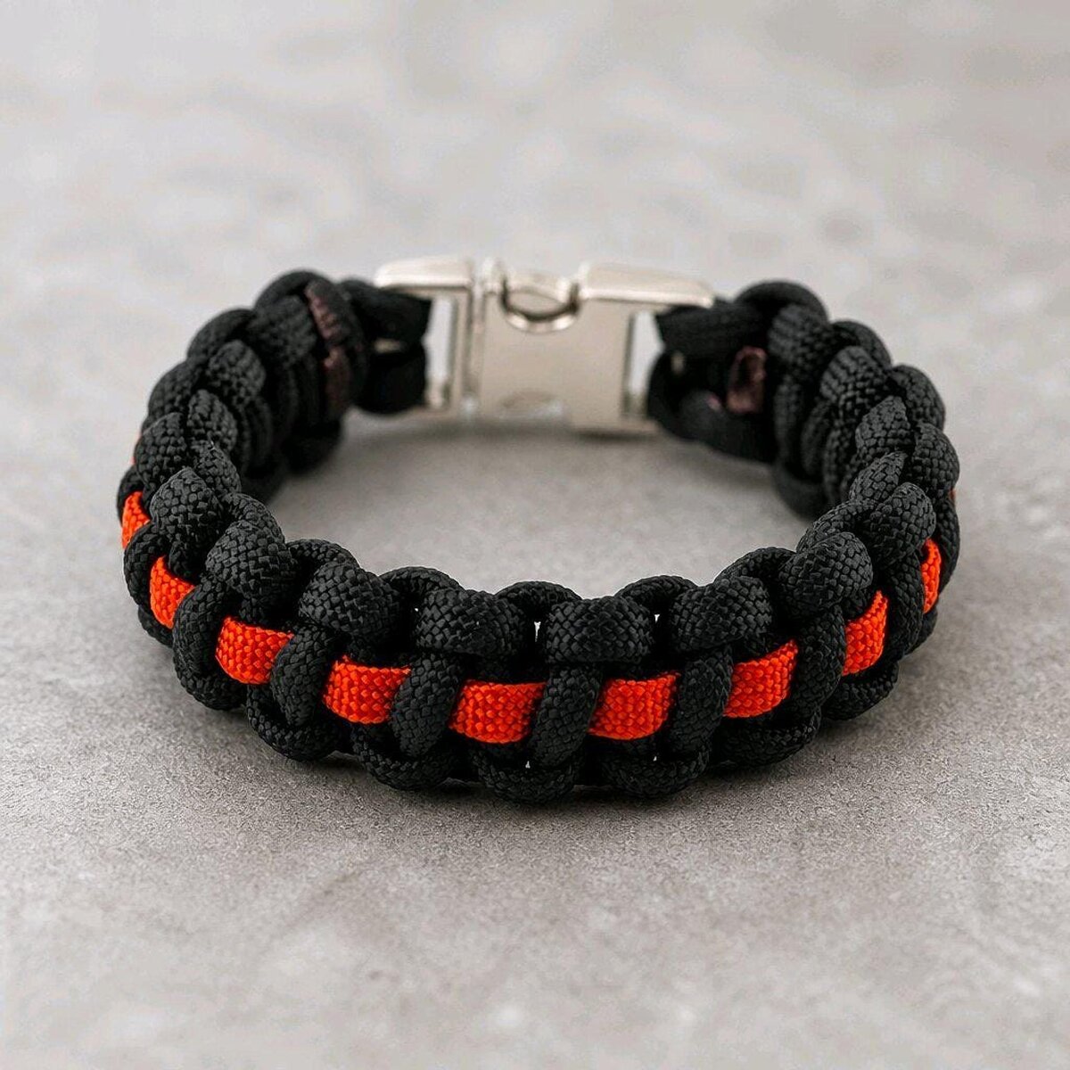 A black and bright orange paracord bracelet with a silver side-release buckle, lying on a light grey textured surface.