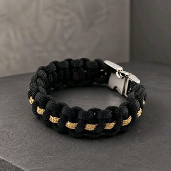 An angled top-down view of the black and gold paracord bracelet with a connected silver buckle on a gray textured background.