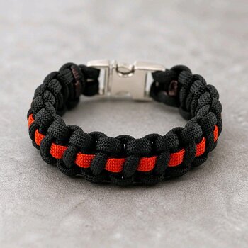 A black and bright orange paracord bracelet with a silver side-release buckle, lying on a light grey textured surface.